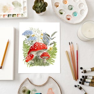 May include: A watercolor painting of red and white mushrooms with blue and yellow flowers and green ferns. The painting is on a white background and is surrounded by art supplies, including paint palettes, brushes, and paint tubes.