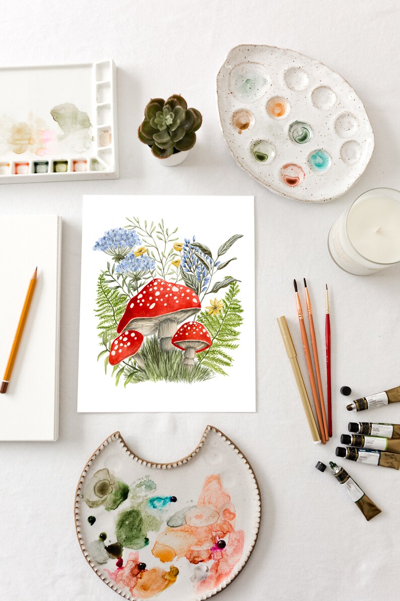 May include: A watercolor painting of red and white mushrooms with blue and yellow flowers and green ferns. The painting is on a white background and is surrounded by art supplies, including paint palettes, brushes, and paint tubes.