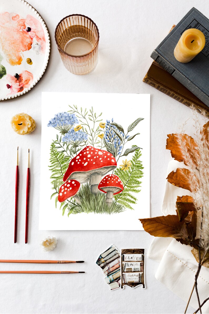 May include: Watercolor painting of red and white mushrooms with white spots growing in a cluster of green ferns and blue and yellow wildflowers.