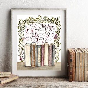 May include: A framed watercolor print with a quote about books: "Between the pages of a book is a lovely place to be." The quote is surrounded by a wreath of green leaves and pink flowers. A row of vintage books are displayed below the quote.