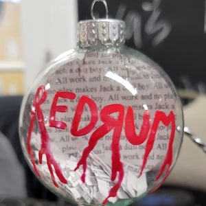 Puede incluir: Un adorno navideño de vidrio transparente con una inscripción roja "REDRUM" y pintura roja que gotea como sangre. El adorno está lleno de papel triturado.
