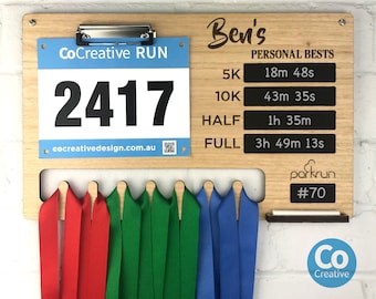 May include: A wooden medal display board with the name "Ben's" and a personalized running time tracker. The board has a number tag with the number "2417" and the text "CoCreative RUN". The board also has a place to hang medals with the text "parkrun #70".