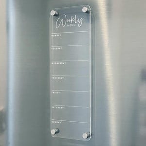 May include: A clear acrylic weekly menu board with a white inscription that reads "Weekly Menu". The board has seven lines for each day of the week, starting with Monday and ending with Sunday.