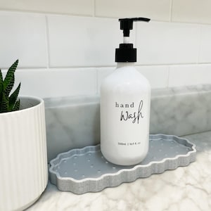 Op de afbeelding: Witte keramische handzeepdispenser met een zwarte pomp en de tekst "hand wash" en "500ml | 16.9 fl. oz" op een grijs gevlekt dienblad.
