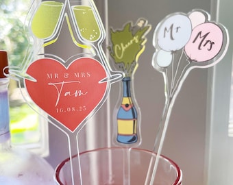 May include: Clear acrylic drink stirrers with wedding-themed designs. One stirrer features champagne glasses and a heart with "Mr & Mrs Tam" and a date. Other stirrers include a champagne bottle with "Cheers" and balloons with "Mr & Mrs".