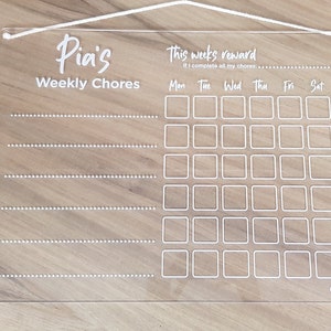 May include: A clear acrylic weekly chore chart with a white string for hanging. The chart has a title that says "Pia's Weekly Chores" and a section for "This week's reward" with the text "If I complete all my chores: ...". The chart has a grid of squares for each day of the week, labeled "Mon", "Tue", "Wed", "Thu", "Fri", "Sat", and "Sun". The bottom right corner of the chart has the text "CoCreative".