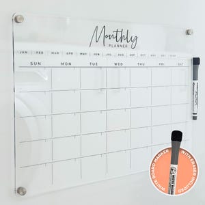 May include: A clear acrylic monthly planner with the words "Monthly Planner" in elegant script. The planner includes a grid for daily scheduling, with months and days of the week labeled. A white board marker with an eraser is included.