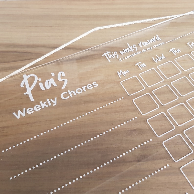 May include: A clear acrylic chore chart with white text that reads "Pia's Weekly Chores". The chart has a section for a weekly reward with the text "This week's reward If I complete all my chores..." and a grid with days of the week labeled "Mon", "Tue", "Wed", "Thu", and "Fri".