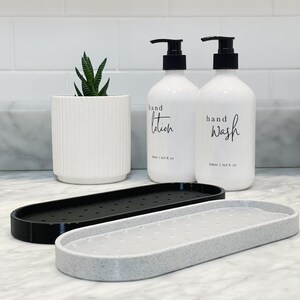 Op de afbeelding: Een moderne badkamerset met twee witte pompflessen met de tekst "hand lotion" en "hand wash", een witte plantenbak met een vetplant en twee zeepschalen. De flessen zijn elk 500 ml.