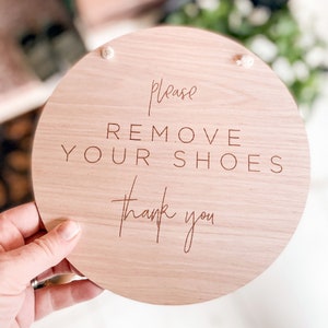 Op de afbeelding: Een rond houten bord met de tekst "Alstublieft schoenen uitdoen Dank u" in lichtbruin.