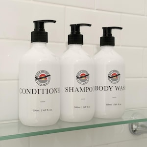 May include: Three white plastic pump bottles labelled Conditioner, Shampoo, and Body Wash. Each bottle features a circular logo with a black aeroplane silhouette over a red and orange sunset, the text Paul & Deborah Est 2025, and 500ml | 16.9 fl. oz.