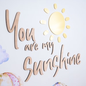 Peut inclure: Décoration murale en bois avec les mots "You are my sunshine" en écriture cursive. Un soleil doré avec des rayons est au-dessus des mots.