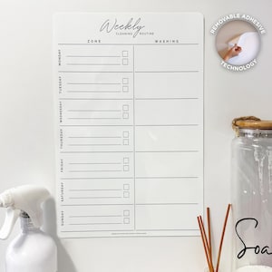 Könnte beinhalten: Eine weiße Wochenplan-Reinigungstafel mit den Worten "Weekly Cleaning Routine" oben. Die Tafel hat Abschnitte für jeden Wochentag mit Kontrollkästchen für Zonen- und Waschaufgaben. Der Text "Removable Adhesive Technology" ist ebenfalls sichtbar.