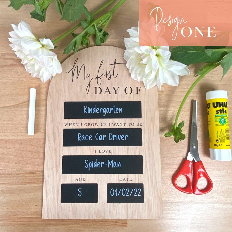 Puede incluir: Un letrero de madera con un inserto de pizarra para el primer d&iacute;a de jard&iacute;n de infantes de un ni&ntilde;o. El letrero tiene el texto "My first day of" e incluye espacios para el grado del ni&ntilde;o, lo que quiere ser cuando sea grande, lo que le gusta, su edad y la fecha. El letrero est&aacute; decorado con flores y un pegamento.