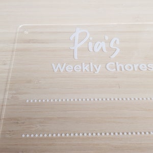 May include: A clear acrylic sign with the text "Pia's Weekly Chores" in white lettering. The sign has two rows of small white dots.