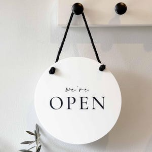 May include: A circular white sign with the black text "We're OPEN" printed in the center. The sign is suspended by a black braided rope that is knotted through two holes at the top, allowing it to hang from a hook on a white wall surface.