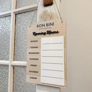 May include: A rectangular light wood sign with a silver chain for hanging. It features the text BON BINI AESTHETICS and Opening Hours in black. A white acrylic panel with horizontal black lines lists the days from Monday to Sunday for writing.