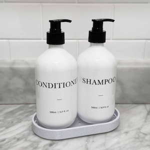 May include: Two white plastic bottles with black pump dispensers, labeled CONDITIONER and SHAMPOO in black text. Each bottle displays 500ml | 16.9 fl. oz. The bottles sit on a light gray oval tray. The design is clean, functional, and minimalist.