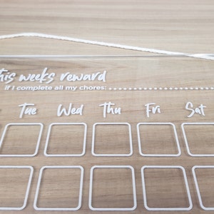 May include: A clear acrylic weekly reward chart with white text and a white string for hanging. The chart has a section for each day of the week, with boxes for marking off chores completed. The text reads "This week's reward if I complete all my chores: Mon Tue Wed Thu Fri Sat Sun"