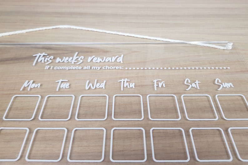 May include: A clear acrylic weekly reward chart with white text and a white string for hanging. The chart has a section for each day of the week, with boxes for marking off chores completed. The text reads "This week's reward if I complete all my chores: Mon Tue Wed Thu Fri Sat Sun"
