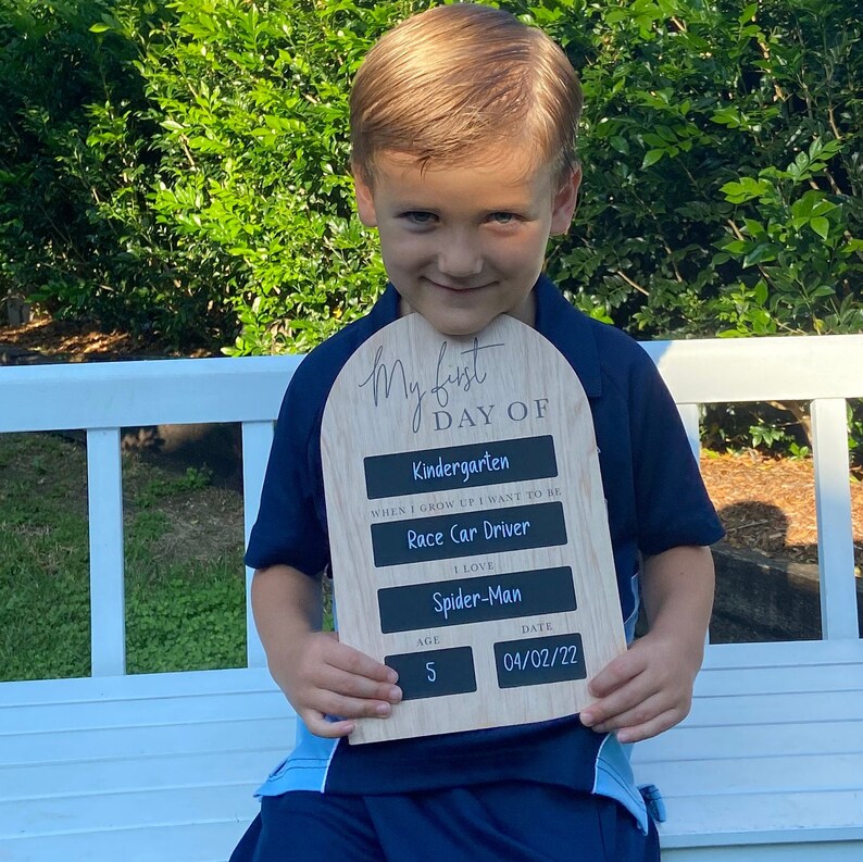 Puede incluir: Un ni&ntilde;o sostiene un letrero de madera que dice "My First Day Of Kindergarten". El letrero tambi&eacute;n incluye la edad del ni&ntilde;o, 5 a&ntilde;os, y la fecha, 04/02/22. El letrero tiene un fondo de pizarra y al ni&ntilde;o le gusta "Spider-Man".