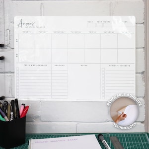 Puede incluir: Un planificador de estudio blanco con el texto "Angus Study Planner" en la parte superior. El planificador tiene secciones para cada día de la semana, con espacios para pruebas, tareas, plazos, notas y temas. Está montado en una pared de ladrillo blanco.