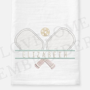 Könnte beinhalten: Weißes Leinenhandtuch mit einem gestickten Tennis-Schläger- und Ball-Design. Das Design umfasst zwei Schläger, einen Tennisball und den Namen "ELIZABETH" in hellgrüner Schrift. Die Schläger haben eine hellgraue Umrandung und einen beigen Griff.