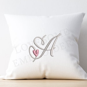 May include: A white square pillow with a large embroidered letter "A" in a cursive font, accompanied by a pink heart. The pillow is on a light wooden surface.