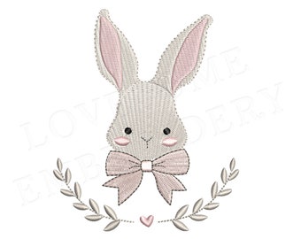 May include: An embroidery design featuring a light gray rabbit head with long ears, pink inner ears, black eyes, and pink cheeks. A light pink bow is positioned below the chin, framed by two curved leafy branches and a small pink heart at the bottom.