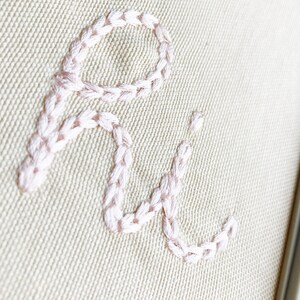 May include: Close-up of a beige fabric with the letters "Ri" embroidered in pink thread.