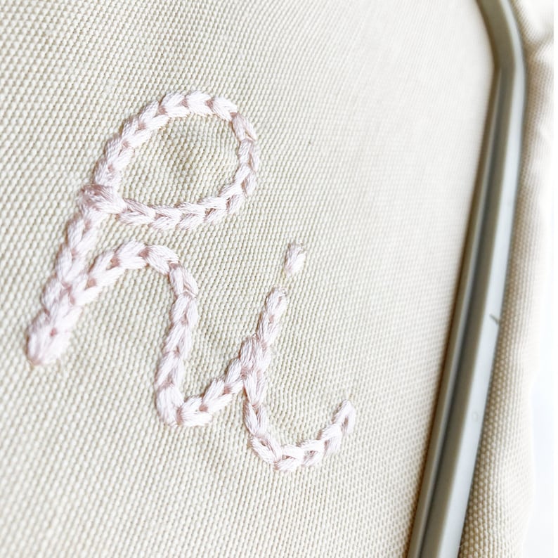 May include: Close-up of a beige fabric with the letters "Ri" embroidered in pink thread.