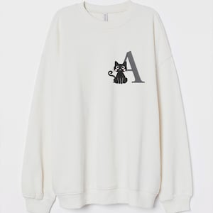May include: White sweatshirt with a black cat and the letter A embroidered on the front.