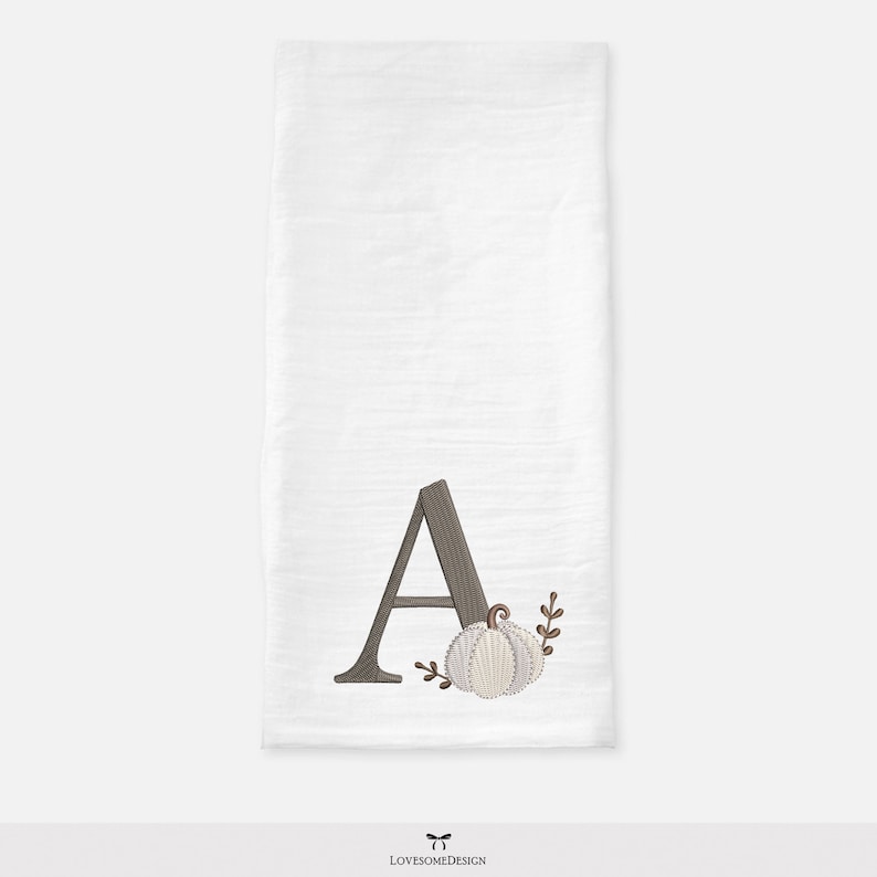 Peut inclure: Un torchon de cuisine rectangulaire blanc orn&eacute; d'une broderie repr&eacute;sentant la lettre majuscule A avec un motif de grain de bois brun fonc&eacute;, accompagn&eacute;e d'une petite citrouille claire et de deux fines branches brunes sans feuilles.