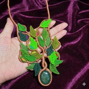 May include: A handmade macrame necklace featuring a tree design with green leaves and a brown trunk. The necklace has a large oval green stone pendant. The leaves have accents of yellow, pink, and blue.