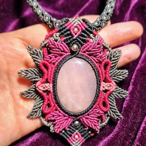 May include: A macrame necklace featuring a large, oval, pale pink rose quartz cabochon at the center. The stone is surrounded by intricate knotwork in black, dark gray, and vibrant magenta thread, accented with small, polished silver-toned metal beads.