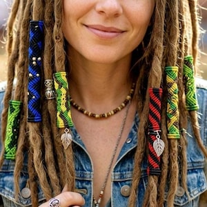 May include: A collection of colorful woven hair wraps for dreadlocks, featuring geometric patterns in blue, green, yellow, and red. Some wraps include silver-toned metal leaf charms and small metallic beads, worn on brown dreadlocks.