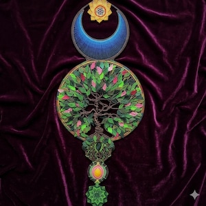 Puede incluir: Un tapiz de pared con cuentas que presenta un diseño de árbol de la vida. La obra de arte incluye una luna creciente azul, una estrella dorada y un árbol circular con hojas verdes y rosas. La pieza está sobre un fondo de terciopelo morado oscuro.