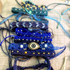 Può includere: Una collezione di braccialetti in macramè fatti a mano in varie tonalità di blu, caratterizzati da intricati motivi intrecciati, perline metalliche e un ciondolo centrale a forma di occhio con un design realistico dell'iride.