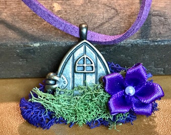 May include: A silver charm necklace with a small house charm, a purple flower, and green moss. The necklace is on a purple suede cord.