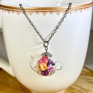 May include: A clear glass teapot pendant necklace filled with colorful dried flowers. The pendant hangs from a silver-colored chain. The necklace is displayed in front of a white teacup with gold trim.