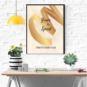May include: A framed art print with a black border. The artwork features gold brushstrokes and the words "Grace for Speed" with "PROVERBS 4:12" below. The print is displayed on a wooden table with decorative items.