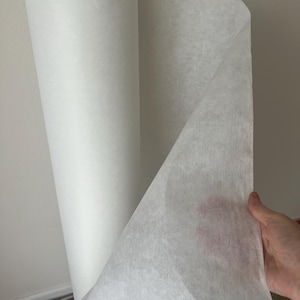 May include: A roll of white interfacing fabric, used for stabilizing and strengthening fabric, is partially unrolled and held in a hand.