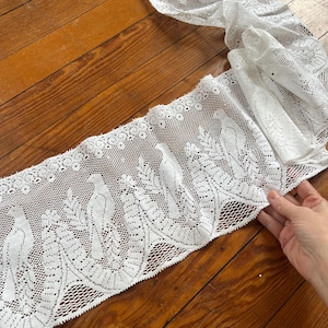 May include: White lace trim featuring a repeating pattern of birds perched on branches. The delicate fabric is displayed on a wooden surface, showcasing the intricate openwork design. This trim is suitable for various craft and sewing projects.