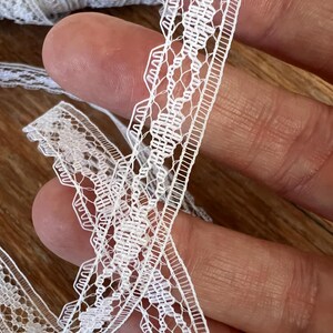 May include: Close-up of white lace trim with a delicate, openwork design. The lace features a scalloped edge and a repeating geometric pattern. A roll of the same lace is visible in the background, suggesting a craft or sewing supply.