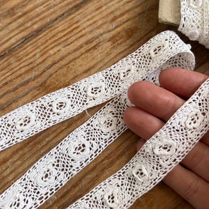 May include: Close-up of white lace trim with a floral design. The delicate lace features a repeating pattern of small, openwork flowers. The trim is displayed on a wooden surface, showcasing its intricate details and texture. Ideal for sewing and craft projects.