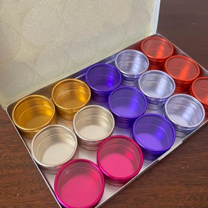 May include: A rectangular metal container filled with small, round, colorful tins. The tins are in various colors, including gold, silver, purple, pink, and orange. The container is open, revealing the arrangement of the tins.