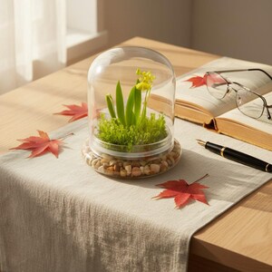 K&ouml;nnte beinhalten: Ein Glasterrarium mit einer transparenten Kuppel, das einen Miniaturgarten mit gr&uuml;nen Pflanzen und gelben Bl&uuml;ten zeigt. Die Basis ist mit kleinen Kieselsteinen gef&uuml;llt. Rote Ahornbl&auml;tter und ein Buch mit Brille und Stift liegen auf dem Tisch.
