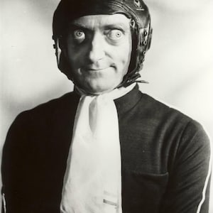 May include: Black and white photo of a man wearing a leather aviator helmet and a white scarf. The man is looking at the camera with a serious expression. The text "BYGONE ERA" is written in gold letters at the bottom of the image.