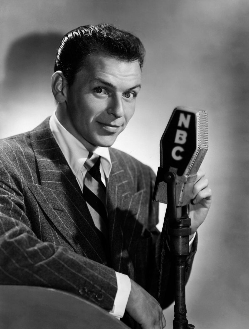 May include: A man in a pinstripe suit and a striped tie is speaking into a microphone with an NBC logo. The microphone is on a stand.