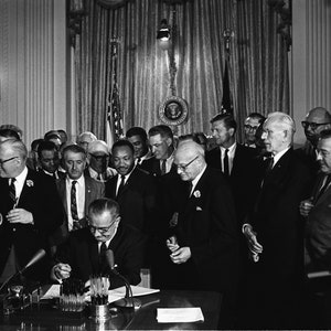 May include: A black and white photo of a group of people in suits gathered around a desk. A man in a suit is signing a document at the desk. The photo was taken in a room with white walls and a large window behind the desk.